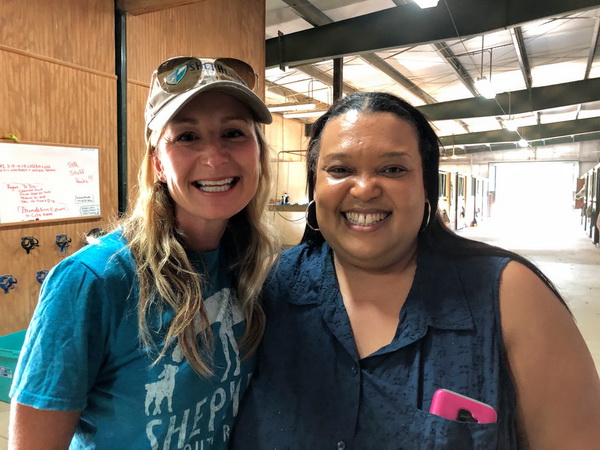 Two women standing in a horse stable are smiling. The person on the left is wearing a hat and light blue shirt. The person on the right is wearing a dark blue top.