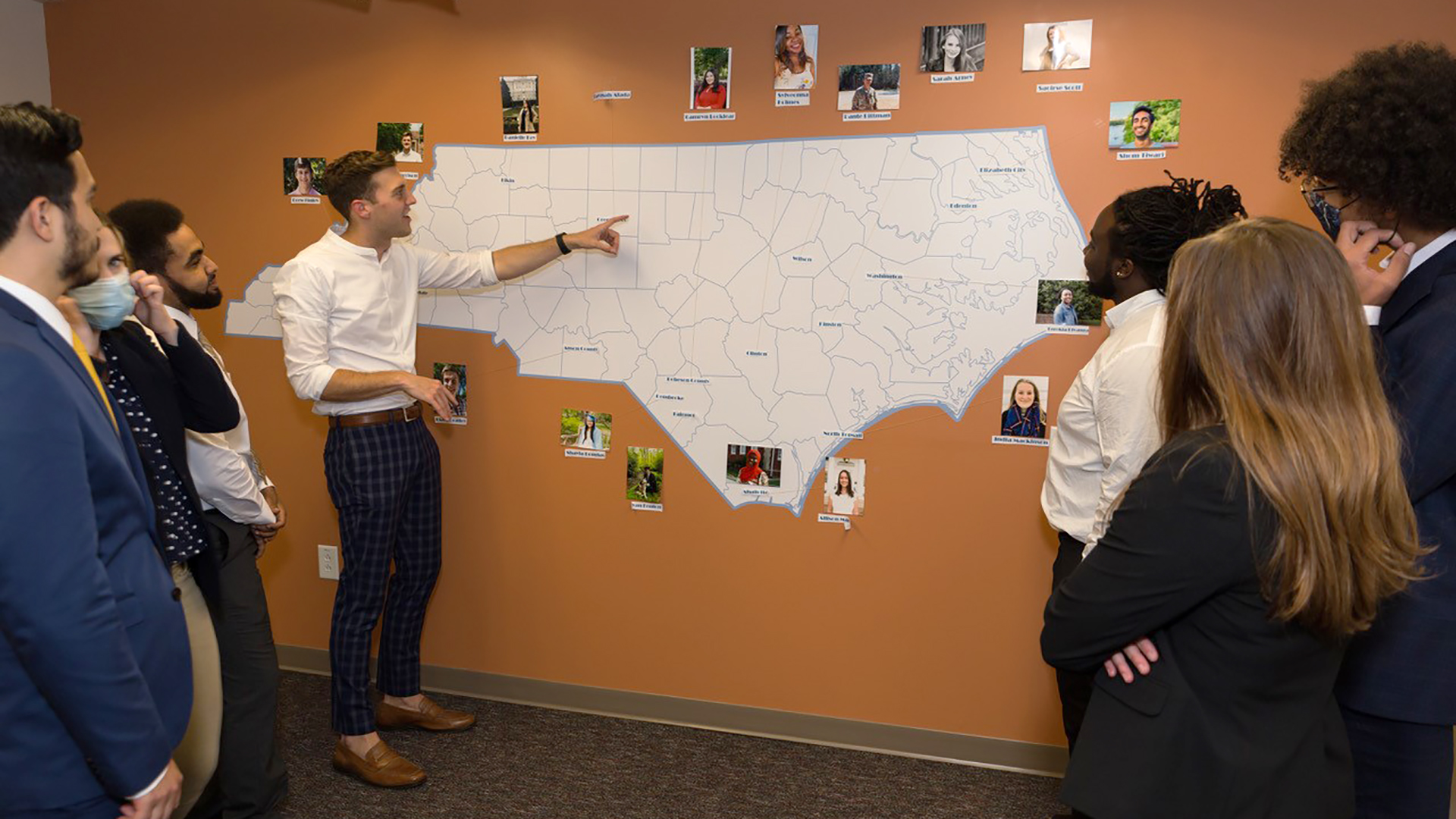 A group of people in business wear gather around a large white map of North Carolina on an orange wall with many photos around the edges.