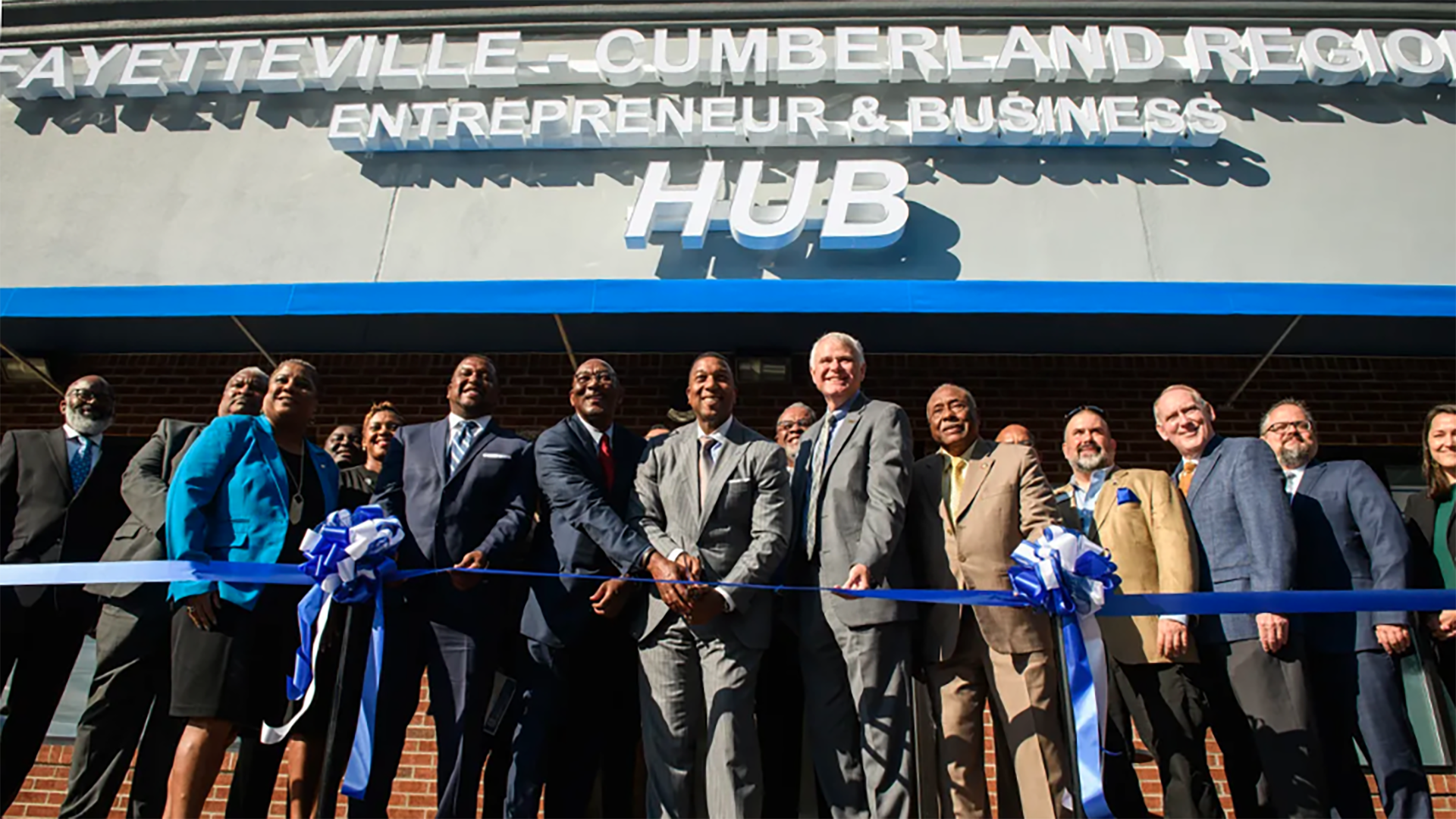 A group of people in suits are cutting a blue ribbon in front of a building that has the words Fayetteville-Cumberland Regional Entrepreneur and Business hub