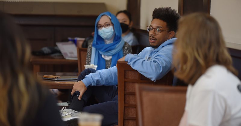 A student wearing a UNC blue sweater talks as a student with long hair and white shirt and a student with a blue head wrap looks on.