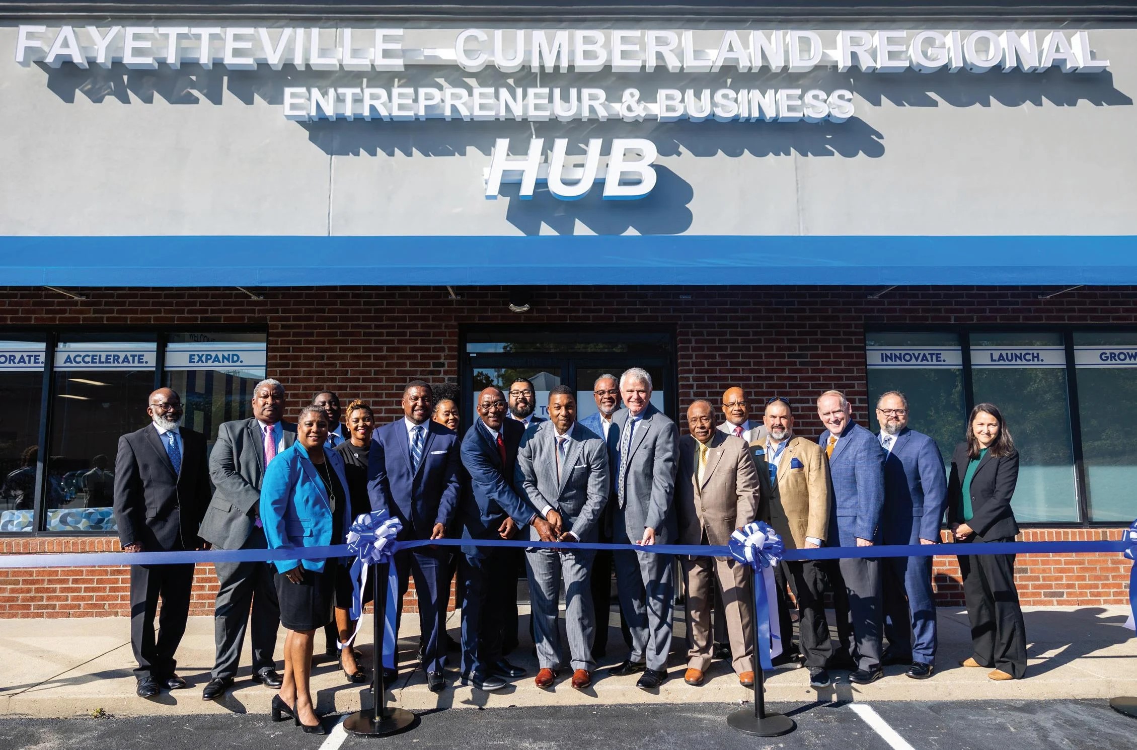 18 individuals in business attire are at a ribbon cutting ceremony. They are standing infront of a building with a sign that says "Fayetteville - Cumberland Regional Entrepreneur & Business Hub."