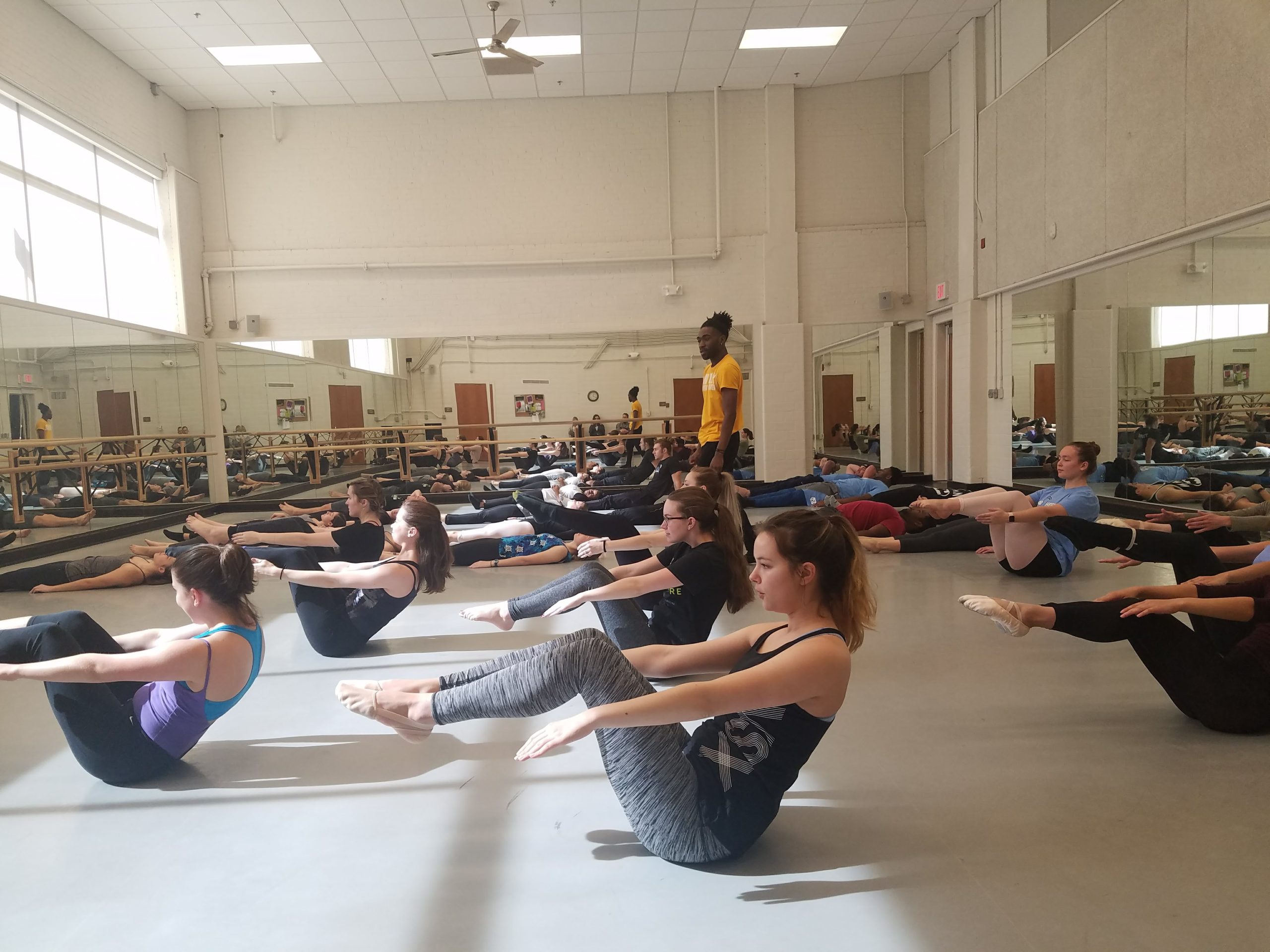 A picture of an instructor standing up and a room full of students in a dance class on the floor doing exercises